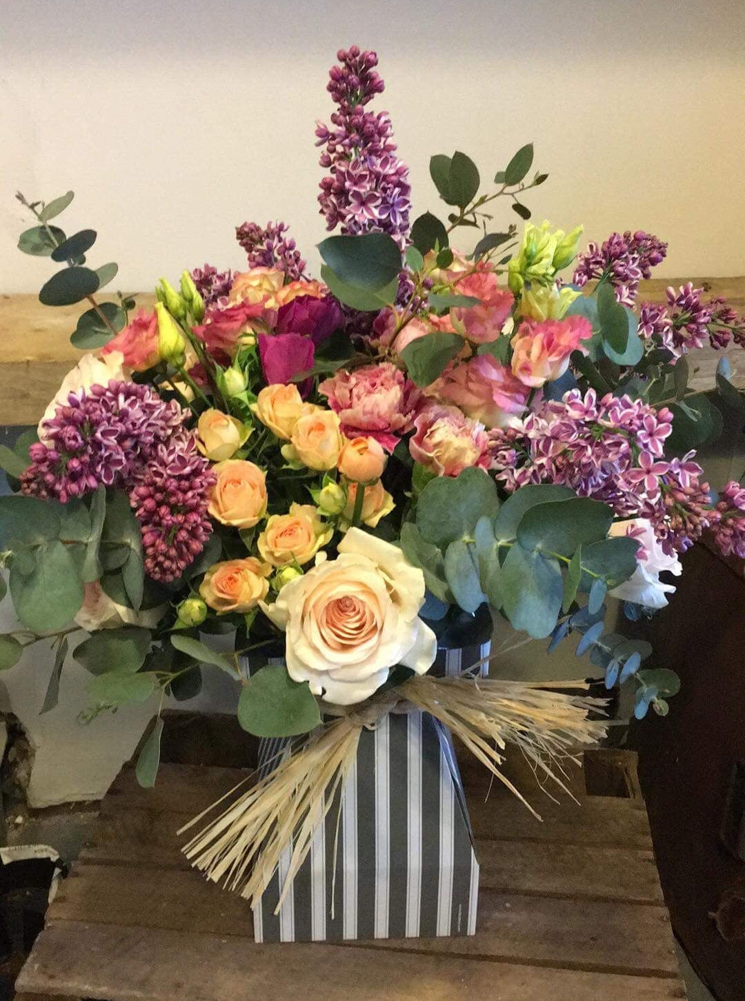 A colorful bouquet of flowers including roses and lilacs, arranged in a striped vase on a wooden surface.