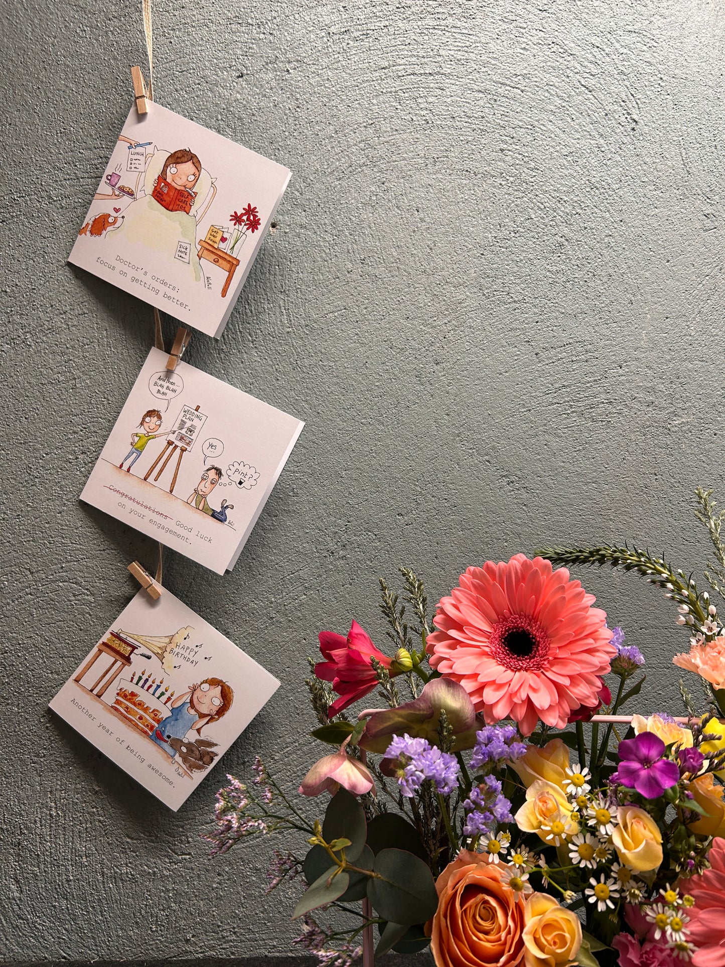 Three small books or cards with illustrations on a string, accompanied by a bouquet of flowers against a gray background.
